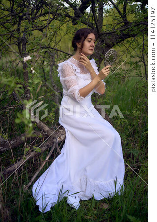 beautiful woman in white vintage dress with train and holding vintage mirror in spring garden at sunset 114125197