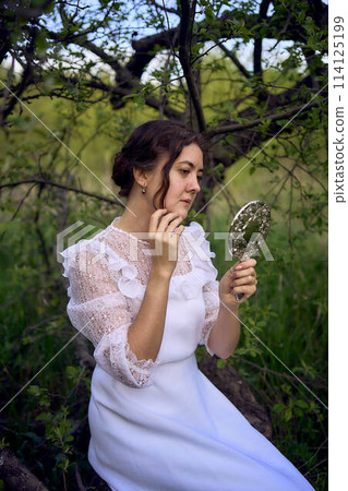 beautiful woman in white vintage dress with train and holding vintage mirror in spring garden at sunset 114125199