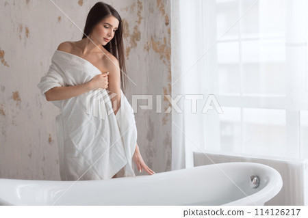 Brown-haired woman in bathrobe, posing with bare shoulders next to bath tube 114126217