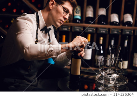 Bartender opening wine bottle with corkscrew in restaurant with wine shelves. Bartender opening wine bottle with corkscrew in restaurant with wine shelves. 114126304