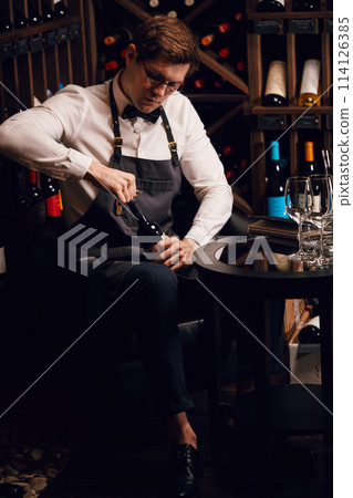 Bartender opening a wine bottle with corkscrew in restaurant with wine shelves. Bartender opening a wine bottle with corkscrew in restaurant with wine shelves. 114126385