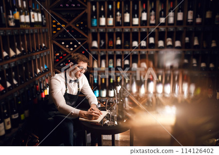 Elegant wine seller holding a bottle of wine and reading label in a wine store Elegant wine seller holding a bottle of wine and reading label in a wine store 114126402