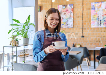 Young woman worker, owner in apron holding cup of coffee in restaurant, coffee shop Young woman worker, owner in apron holding cup of coffee in restaurant, coffee shop 114126918