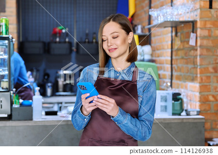 Young woman service worker in apron with smartphone in restaurant, coffee shop Young woman service worker in apron with smartphone in restaurant, coffee shop 114126938