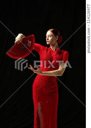 Flamenco female dancer in ornate red dress standing with elegance, posing with red fan against black background Flamenco female dancer in ornate red dress standing with elegance, posing with red fan against black background 114126977