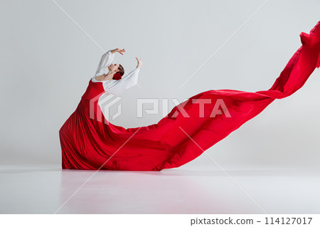 Elegant woman, flamenco dancer in radiant red dress standing in dramatic pose, perfuming against grey studio background. Spirit of dance. 114127017