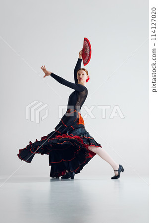 Elegance and emotion. Beautiful artistic woman, performing flamenco dance with classic red fan against grey studio background Elegance and emotion. Beautiful artistic woman, performing flamenco dance with classic red fan against grey studio background 114127020