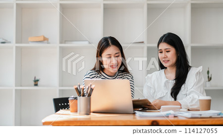 Two colleagues share a light moment while working together at a well-equipped desk, surrounded by an airy and bright office setting. Two colleagues share a light moment while working together at a well-equipped desk, surrounded by an airy and bright office setting. 114127055