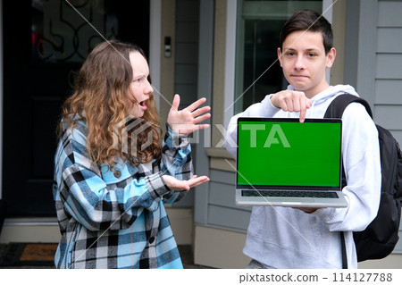 two teenagers in frame boy and girl friend points finger at green chroma key screen on computer laptop girl is surprised with fingers spread open mouth surprised schoolchildren on street advertising 114127788