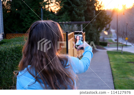 a young woman takes a selfie on street during sunset a teenager girl in her hands with a phone near the house and a fence private sector backlight can be seen reflected in the gadget monitor screen 114127976