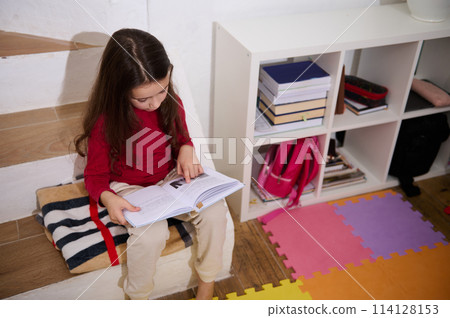 View from above school age child girl reading a book, sitting on steps of her room with multicolored puzzle carpet. The concept of back to school. Smart kids. Erudition and learning. World Book's Day 114128153