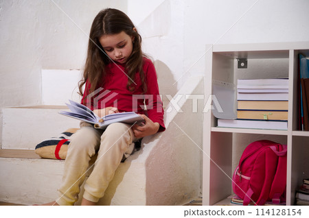 Cute school age child girl reading a book in cozy home atmosphere, sitting on steps of her room. The concept of back to school. Smart kids, Erudition and learning. World Book's Day Cute school age child girl reading a book in cozy home atmosphere, sitting on steps of her room. The concept of back to school. Smart kids, Erudition and learning. World Book's Day 114128154