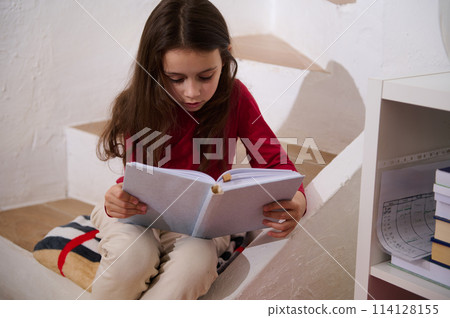 Portrait of cute little school aged girl focused, holding book and reading text, sitting on steps at cozy home interior Portrait of cute little school aged girl focused, holding book and reading text, sitting on steps at cozy home interior 114128155