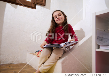 Adorable smart primary school student girl smiling looking at camera, sitting on steps at home with a book in hands. Intelligent little girl reading a book. World Book Day concept Adorable smart primary school student girl smiling looking at camera, sitting on steps at home with a book in hands. Intelligent little girl reading a book. World Book Day concept 114128156