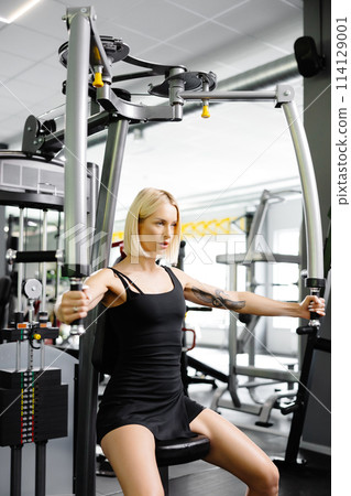 Beautiful and strong athletic woman who using simulator for raising and spreading hands for pumping group of muscles in gym Beautiful and strong athletic woman who using simulator for raising and spreading hands for pumping group of muscles in gym 114129001