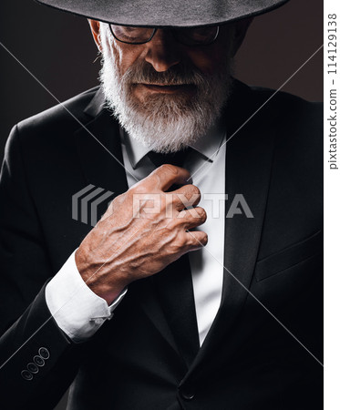 Elderly person hides mournful bearded face behind fedora hat isolated on dark Elderly person hides mournful bearded face behind fedora hat isolated on dark 114129138