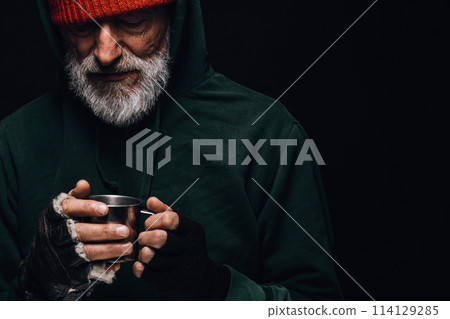 Old homeless man with grey beard holding a mug of hot tea to warm himself. Old homeless man with grey beard holding a mug of hot tea to warm himself. 114129285