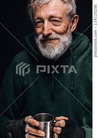 Frozen homeless old man with wrinkled face holds a cup with coins given as alms 114129286