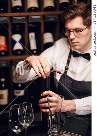 Bartender opening wine bottle with corkscrew in restaurant with wine shelves. Bartender opening wine bottle with corkscrew in restaurant with wine shelves. 114129341