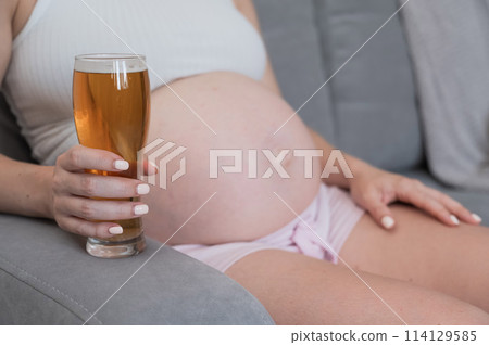 Close-up of the belly of a pregnant woman holding a glass of beer while sitting on the sofa. 114129585