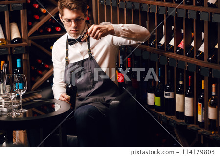 Bartender opening wine bottle with corkscrew in restaurant with wine shelves. Bartender opening wine bottle with corkscrew in restaurant with wine shelves. 114129803