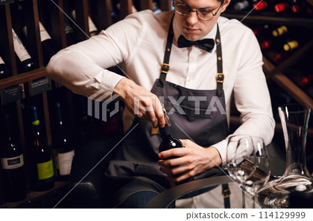 Bartender opening a wine bottle with corkscrew in restaurant with wine shelves. 114129999