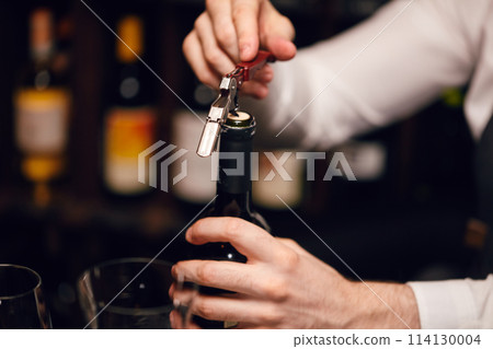 Bartender opening a wine bottle with corkscrew in restaurant with wine shelves. 114130004