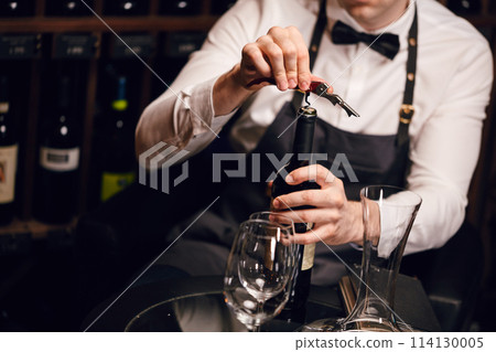Bartender opening a wine bottle with corkscrew in restaurant with wine shelves. 114130005