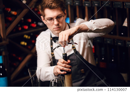 Bartender opening a wine bottle with corkscrew in restaurant with wine shelves. Bartender opening a wine bottle with corkscrew in restaurant with wine shelves. 114130007