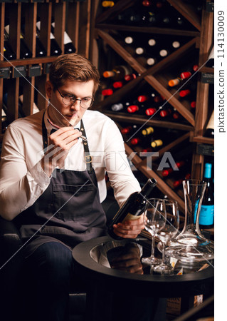 Sommelier opening a wine bottle with corkscrew in restaurant with wine shelves. Sommelier opening a wine bottle with corkscrew in restaurant with wine shelves. 114130009