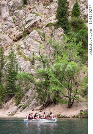 Scenic Mountain Lake with Rocky Cliffs, Forest, Boat, and Tourists on Water 114130274
