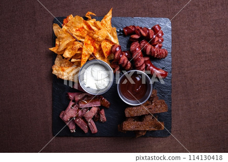 A top-down view of a tantalizing plate featuring bread, savory meat, delicious sausage, crispy chips, and flavorful dip. 114130418