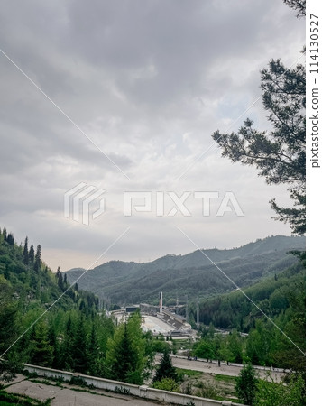 A hydroelectric power plant generates electricity from flowing water. Water channels through a dam and turbine in a valley with mountains. 114130527