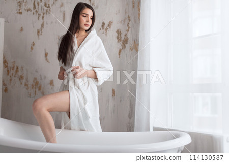 Beautiful young lady in bath robe going to take a bath in white bathroom 114130587