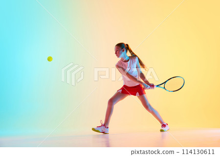 Sportive teen girl, tennis athlete training skills of hitting ball from opponent in neon light against blue-yellow background. 114130611