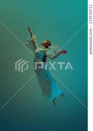 Young woman in white vintage dress exudes sense of deep peace and tranquility, reaching to sky from underwater. 114130711