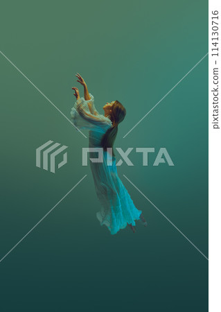 Woman dressed in flowing layers of white extends her arms towards sky from water depth. Model levitating against aqua background. 114130716