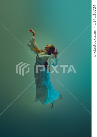 Young woman in cascading white dress reaches upward in graceful motion against serene aqua background. 114130724