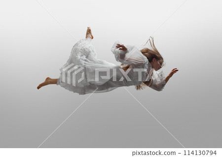 Woman wearing in cloud-like white dress, her hair billowing around her during she levitating in mid-air against white studio background. 114130794