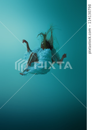 Dynamic photo of young woman floats elegantly, gracefully midair in flowing vintage teal dress against serene aqua background. 114130798