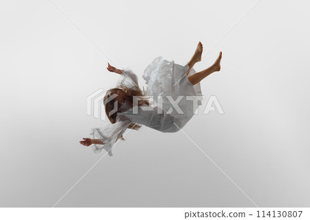 Woman in flowing white dress suspend in air in elegant, graceful pose against white studio background. Dynamic photo. 114130807