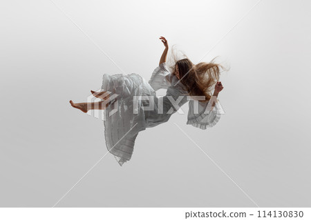 Woman dressed in flowing white gown suspend in mid-air, her long blonde hair cascading around her against white studio background. 114130830