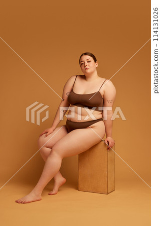 Beautiful young girl with oversized natural body sitting in underwear and posing against light brown studio background Beautiful young girl with oversized natural body sitting in underwear and posing against light brown studio background 114131026