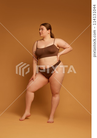 Full-length image of beautiful young girl feeling confident in her natural, plus size body, standing in underwear against light brown studio background Full-length image of beautiful young girl feeling confident in her natural, plus size body, standing in underwear against light brown studio background 114131044