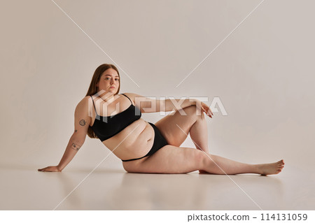 Elegance and confidence. Beautiful young girl with plus size body shape posing on floor in underwear against grey studio background 114131059
