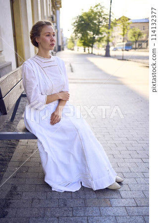 elegant middle age woman in a white vintage dress sitting on a bench in the morning city elegant middle age woman in a white vintage dress sitting on a bench in the morning city 114132377