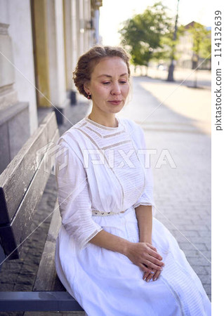 elegant middle age woman in a white vintage dress sitting on a bench in the morning city elegant middle age woman in a white vintage dress sitting on a bench in the morning city 114132639