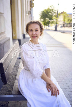 elegant middle age woman in a white vintage dress sitting on a bench in the morning city elegant middle age woman in a white vintage dress sitting on a bench in the morning city 114132641