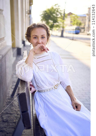 elegant middle age woman in a white vintage dress sitting on a bench in the morning city 114132643