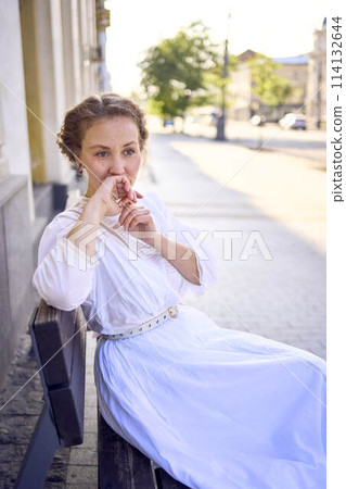 elegant middle age woman in a white vintage dress sitting on a bench in the morning city 114132644